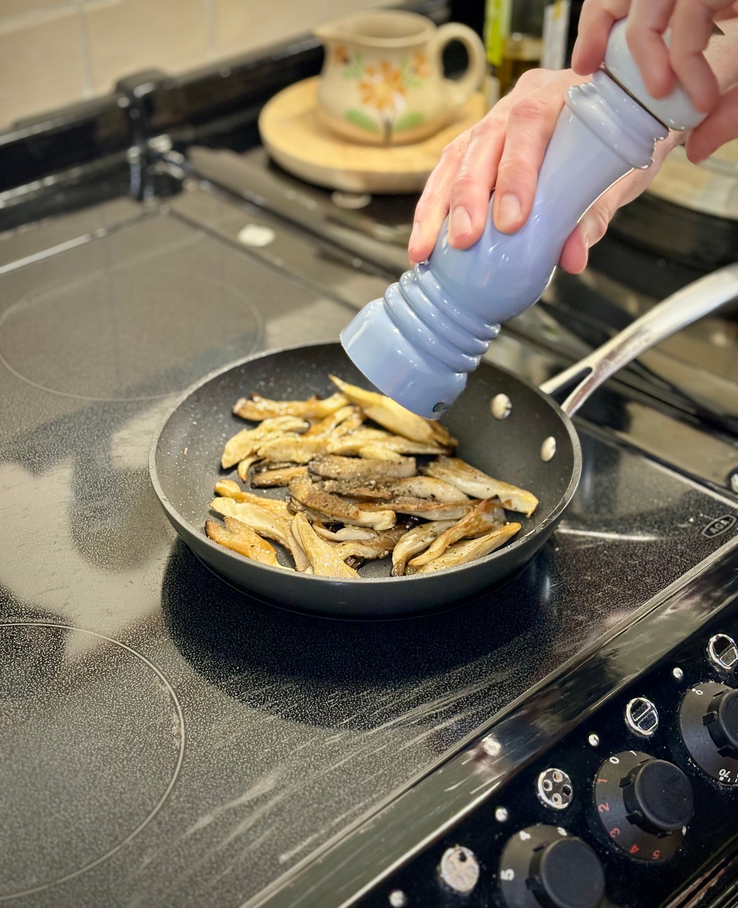 Several shredded blue-grey oyster mushrooms lie in a shallow frying pan. A pair of hands adds cracked black pepper into the pan from a blue pepper grinder. The pan sits on a black induction hob.